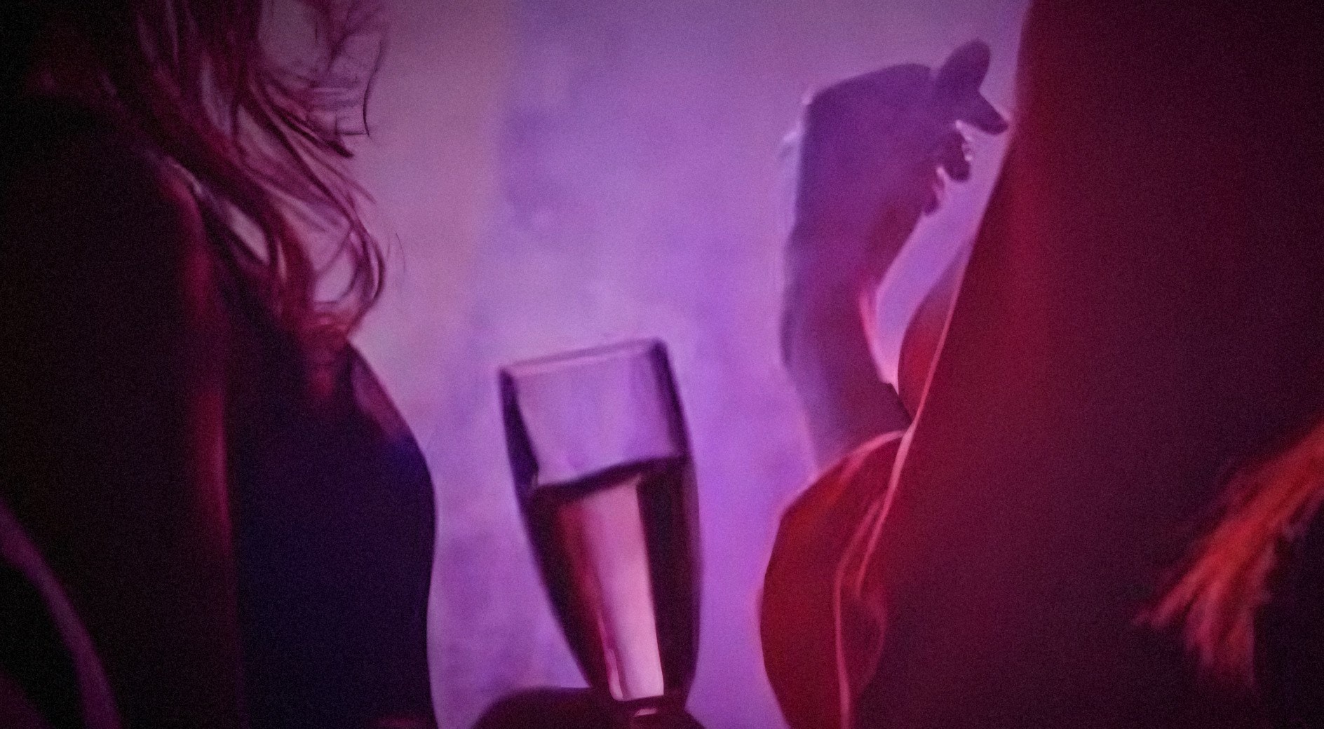 Pessoas de costas sob luz magenta e fumaça, com uma taça de espumante erguida em primeiro plano