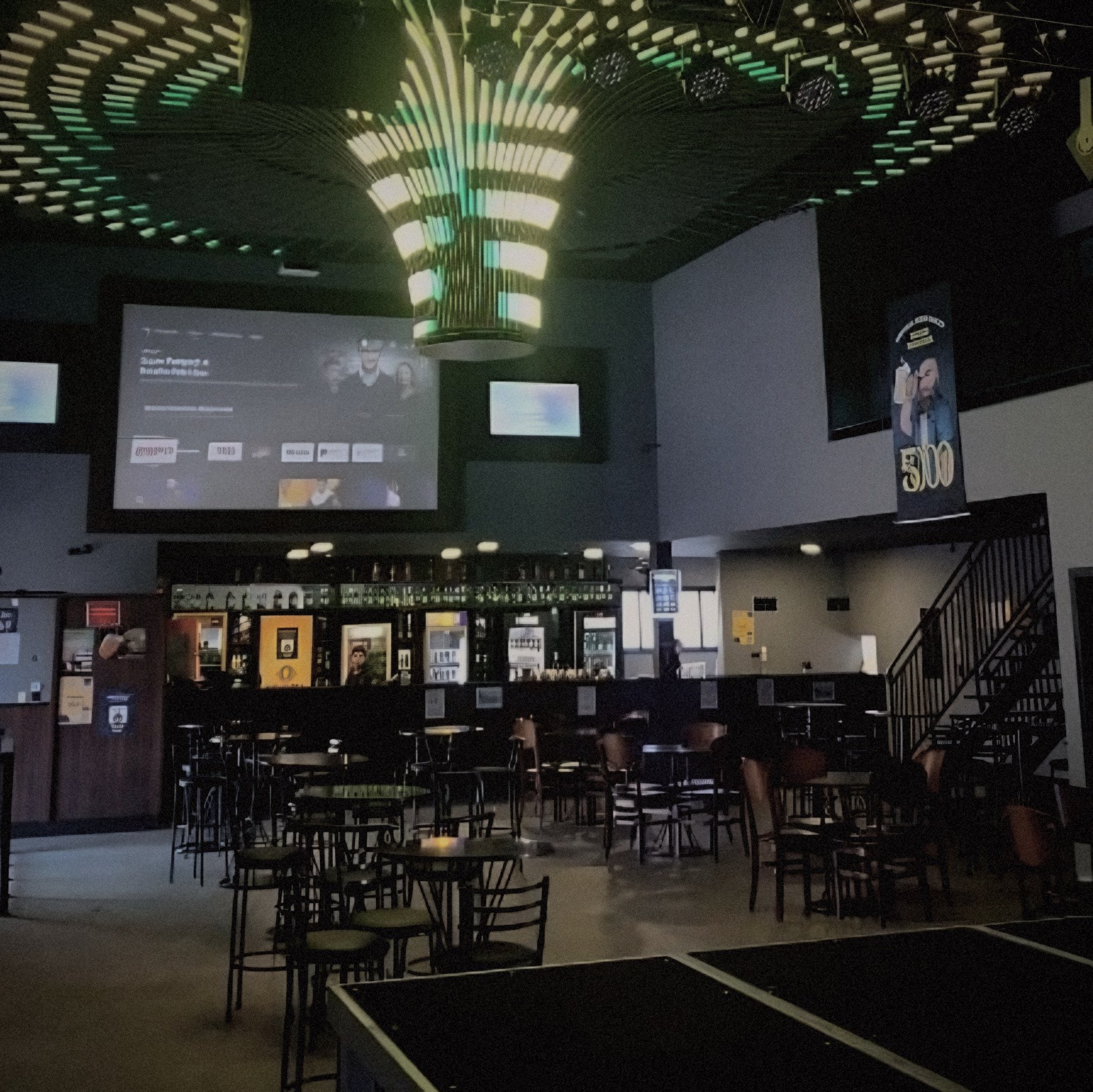 Salão com lustre de LED verde e dourado no teto, telão, balcão do bar ao fundo, mesas altas e escada para o mezanino