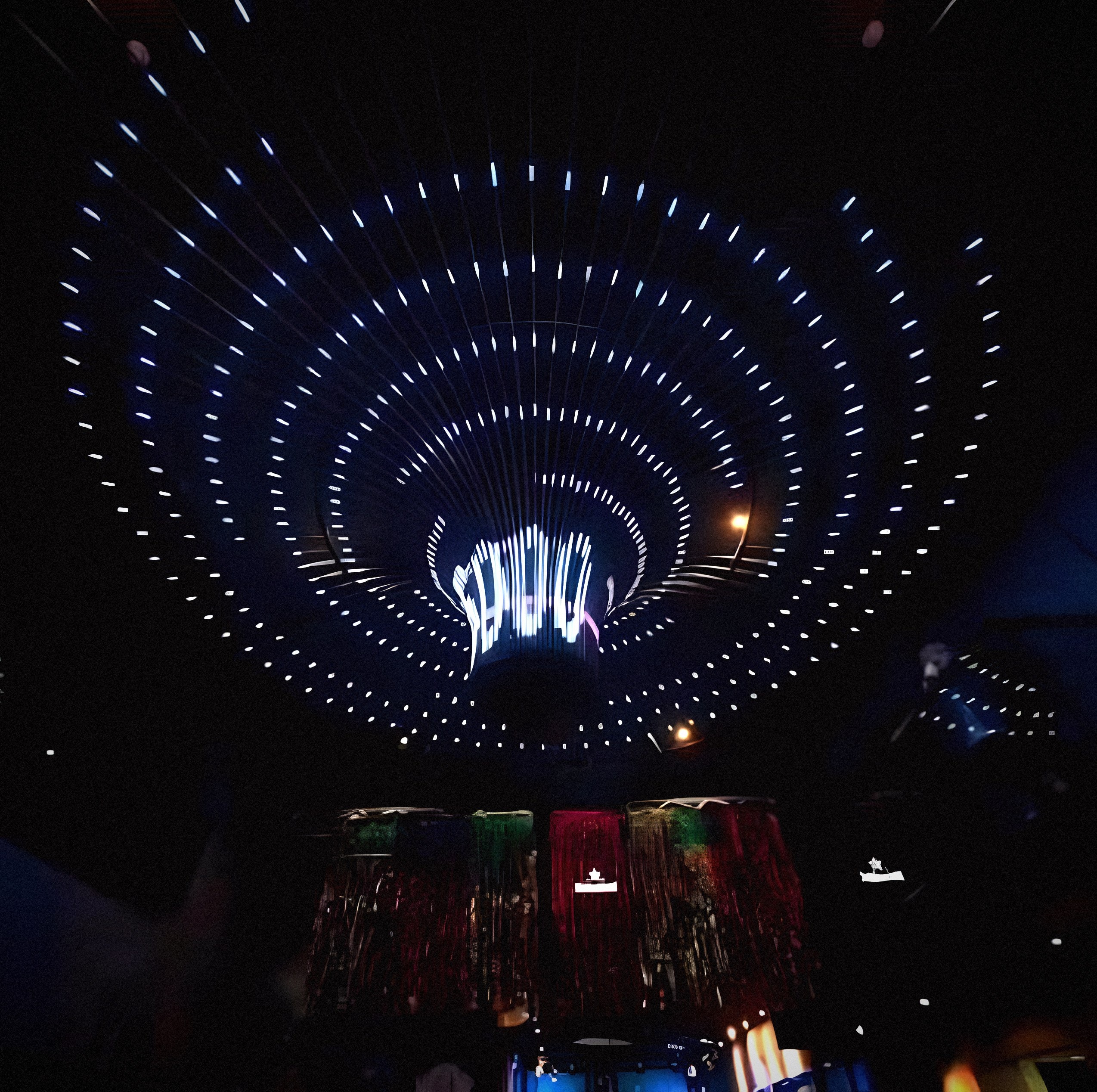 Teto do salão com o lustre de LED circular em anéis de luz e o 5000 aceso no centro, com cortinas de luzes coloridas abaixo